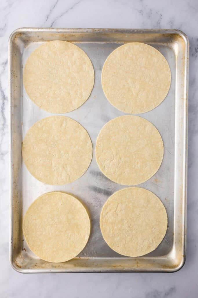 Six corn tortillas are evenly arranged on a metal baking sheet for crispy baked chicken tacos.