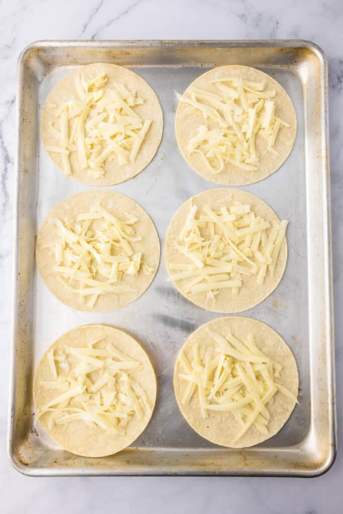 Six corn tortillas topped with shredded cheese are arranged on a metal baking sheet, ready to become crispy baked chicken tacos.