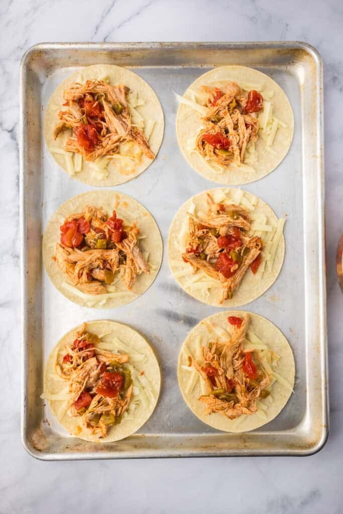 Six corn tortillas on a baking sheet topped with shredded chicken, diced tomatoes, and shredded cheese, ready to be baked into crispy baked chicken tacos.