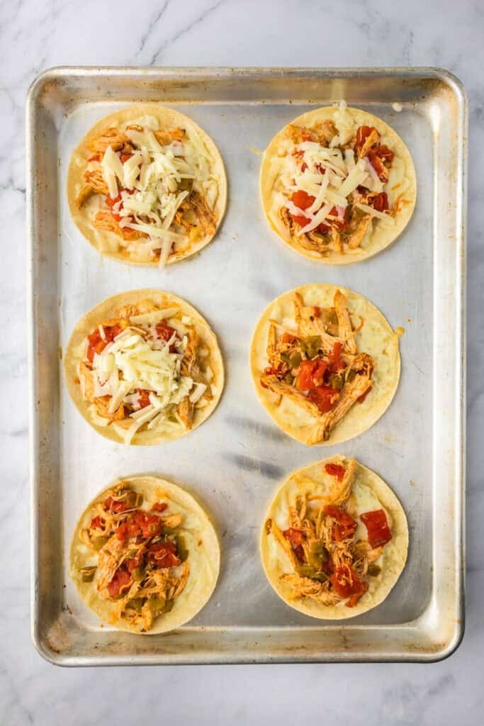 Six corn tortillas topped with shredded chicken, diced tomatoes, chicken, and cheese on a baking sheet, ready to be crispy baked chicken tacos.