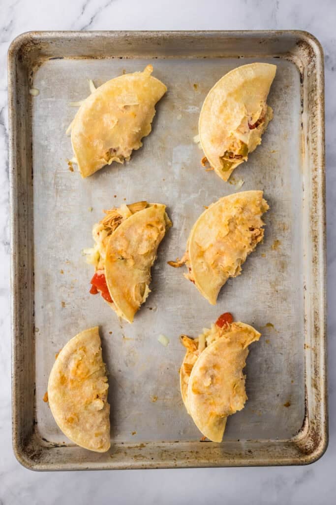 Six folded tacos with visible fillings are arranged on a metal baking sheet. The crispy baked chicken tacos are partially baked with some cheese melted inside.