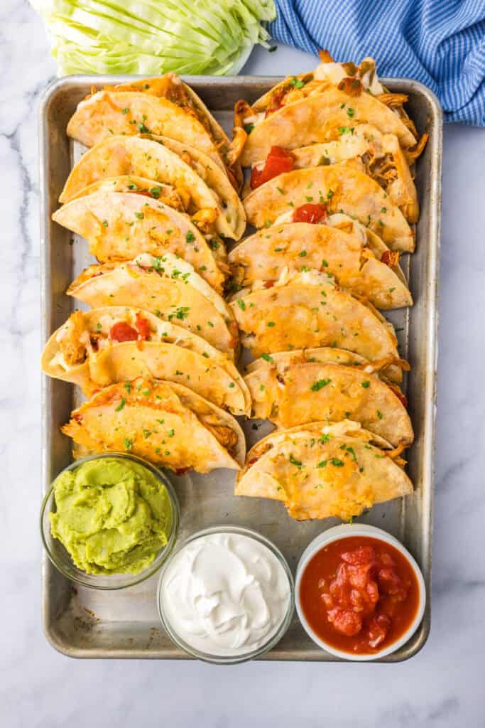 A baking tray with crispy baked chicken tacos topped with melted cheese, served with guacamole, sour cream, and salsa in small bowls.