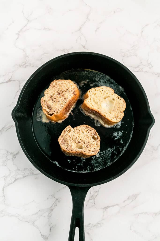 Three slices of french bread being cooked in melted butter in a black cast iron skillet for french bread french toast.