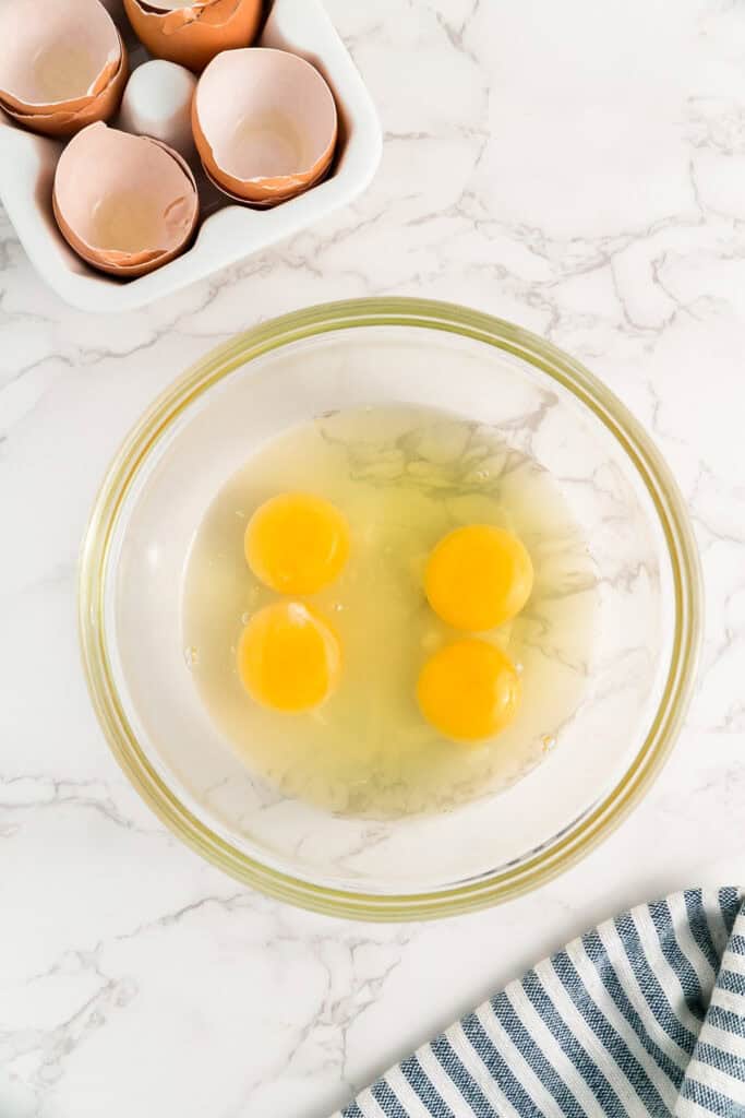 Four raw eggs in a glass bowl for french toast, with empty eggshells on the counter nearby.