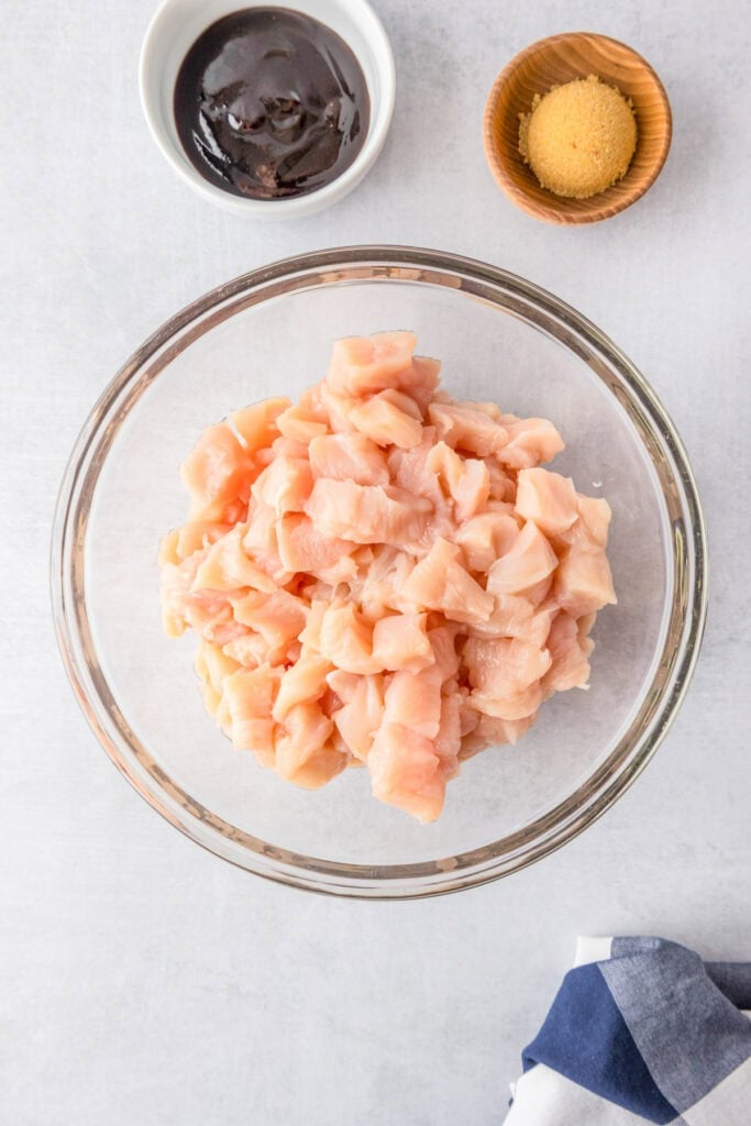 A glass bowl of raw diced chicken with a small bowl of brown sugar and a small bowl of hoison sauce, ready to become Honey Garlic Chicken Noodles.