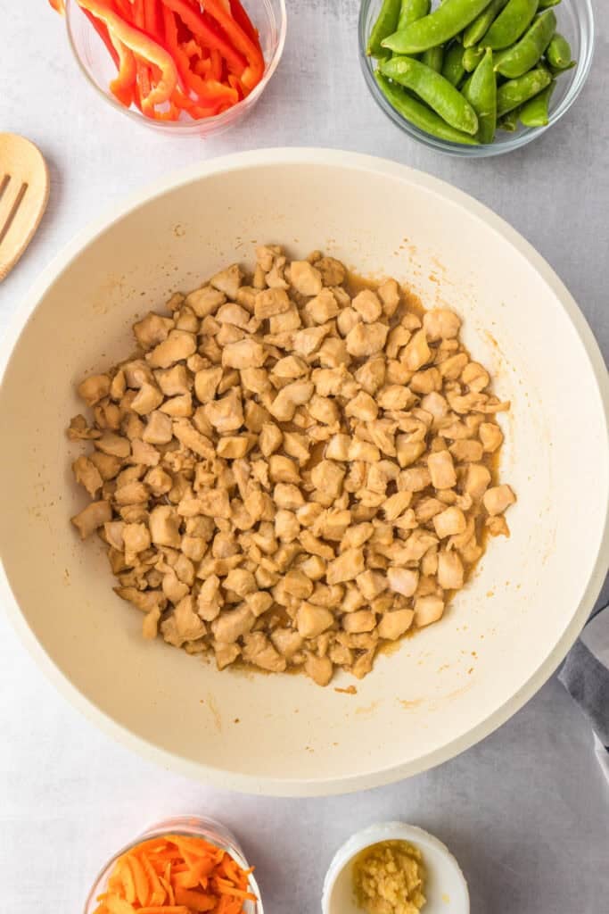 Diced cooked chicken in a pan, surrounded by bowls of sliced red bell peppers, snap peas, shredded carrots, and minced garlic, perfect for making honey garlic chicken noodles.