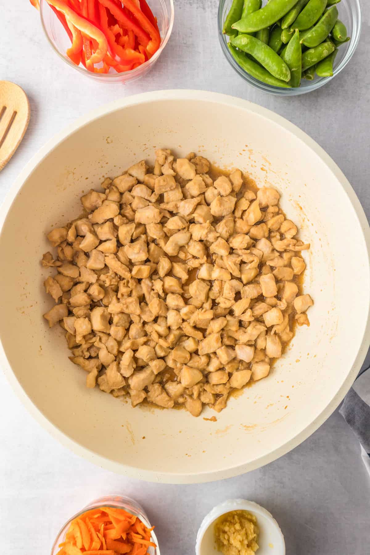 Diced cooked chicken in a pan, surrounded by bowls of sliced red bell peppers, snap peas, shredded carrots, and minced garlic, perfect for making honey garlic chicken noodles.