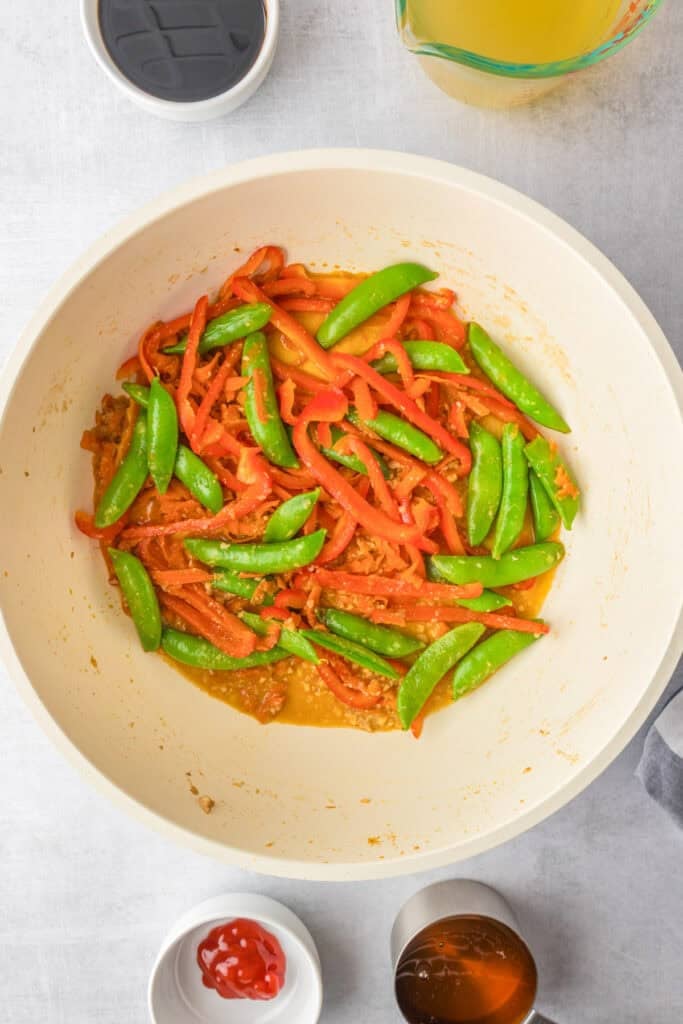 A white pan contains cooked red bell peppers, snap peas, and sliced carrots in a light sauce for honey garlic chicken noodles.
