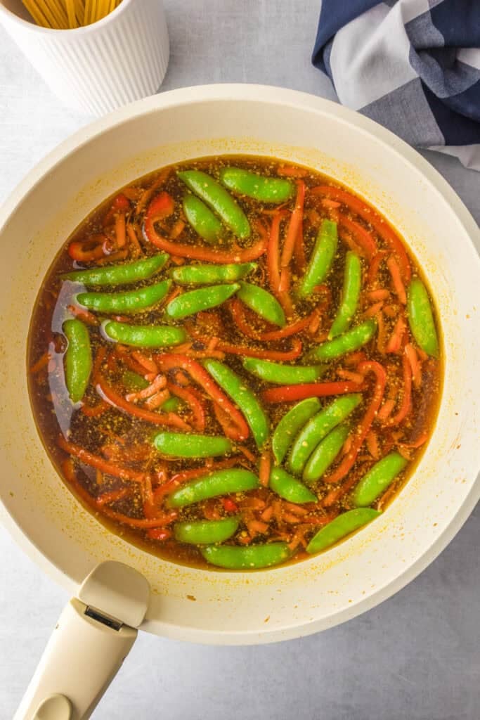 A white skillet containing a broth with sliced red bell peppers, whole snap peas, and uncooked spaghetti nearby for hgoney garlic chicken noodles.