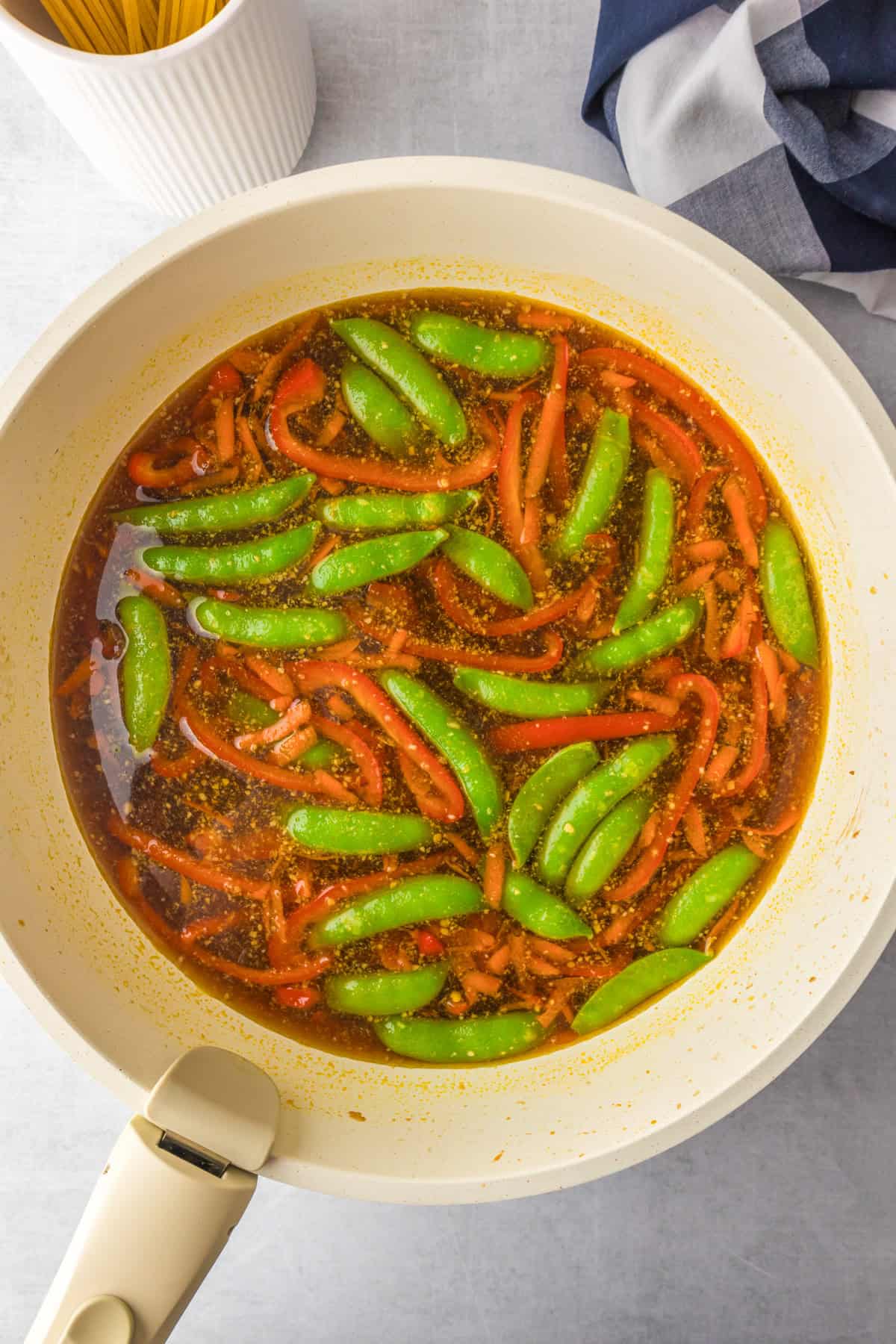 A white skillet containing a broth with sliced red bell peppers, whole snap peas, and uncooked spaghetti nearby for hgoney garlic chicken noodles.