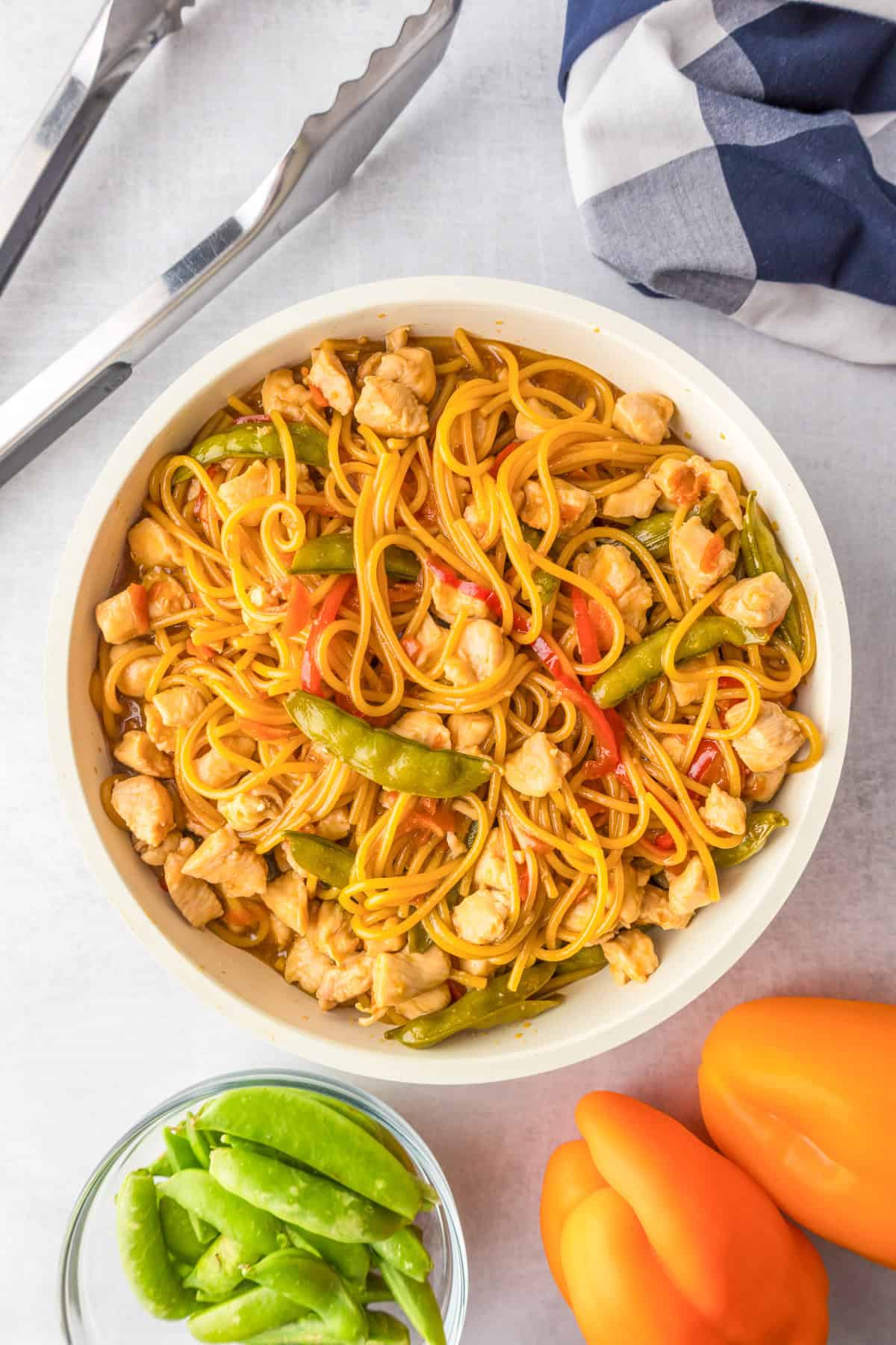 A bowl of honey garlic chicken noodles with bell peppers, snap peas, and sauce, surrounded by metal tongs, a striped napkin, snap peas, and a yellow bell pepper.