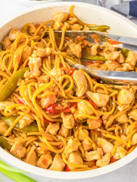 A pan filled with honey garlic chicken noodles, snap peas, and red bell peppers, with a pair of metal tongs resting on the side.
