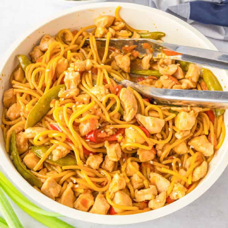 A pan filled with honey garlic chicken noodles, snap peas, and red bell peppers, with a pair of metal tongs resting on the side.
