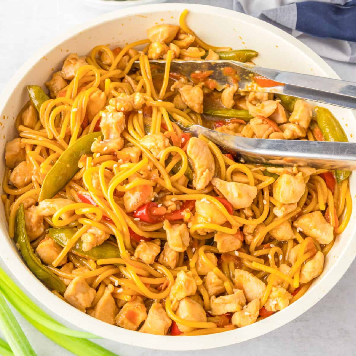 A pan filled with honey garlic chicken noodles, snap peas, and red bell peppers, with a pair of metal tongs resting on the side.