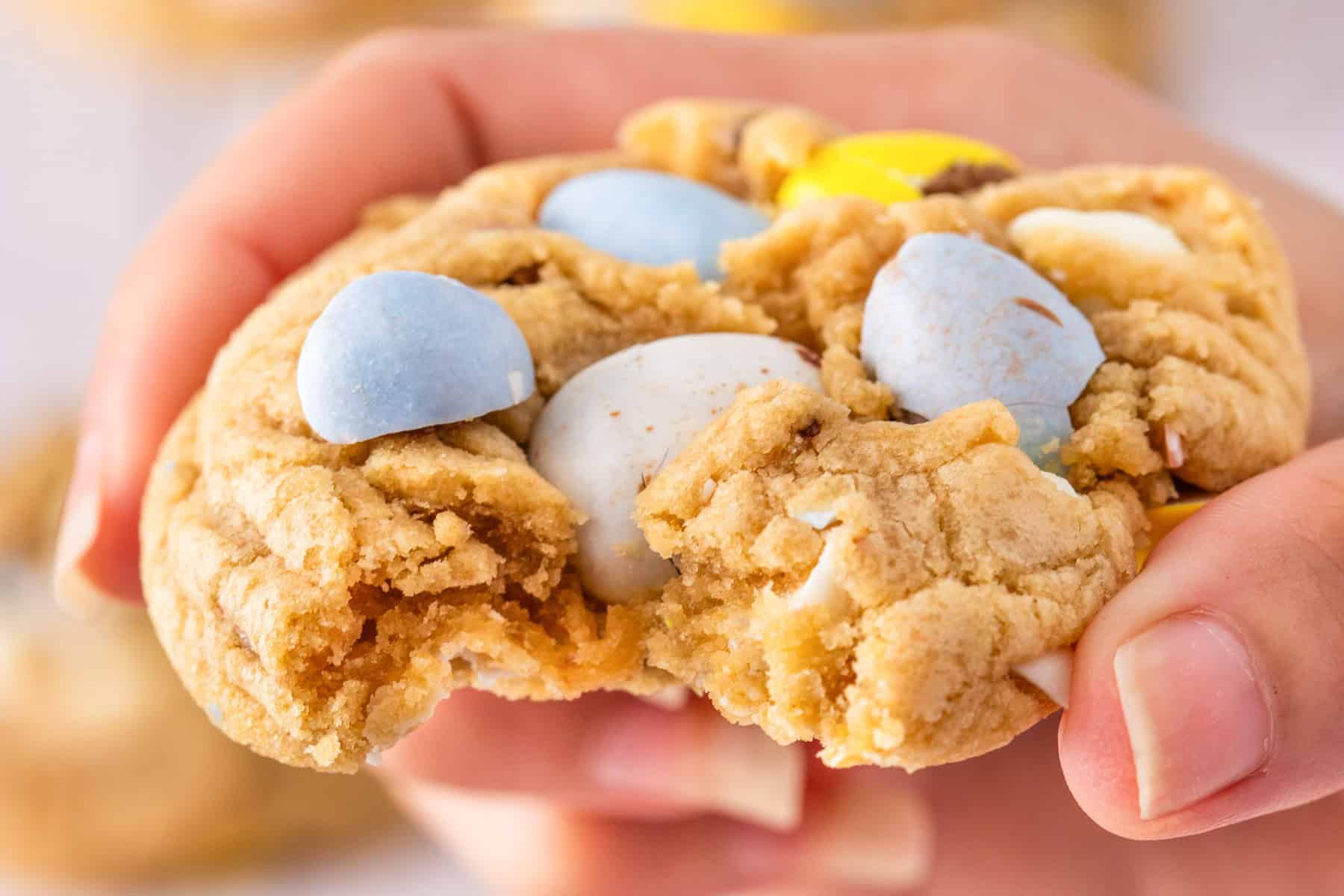 A close-up of a hand holding a partially eaten mini egg cookie, filled with chocolate pieces.