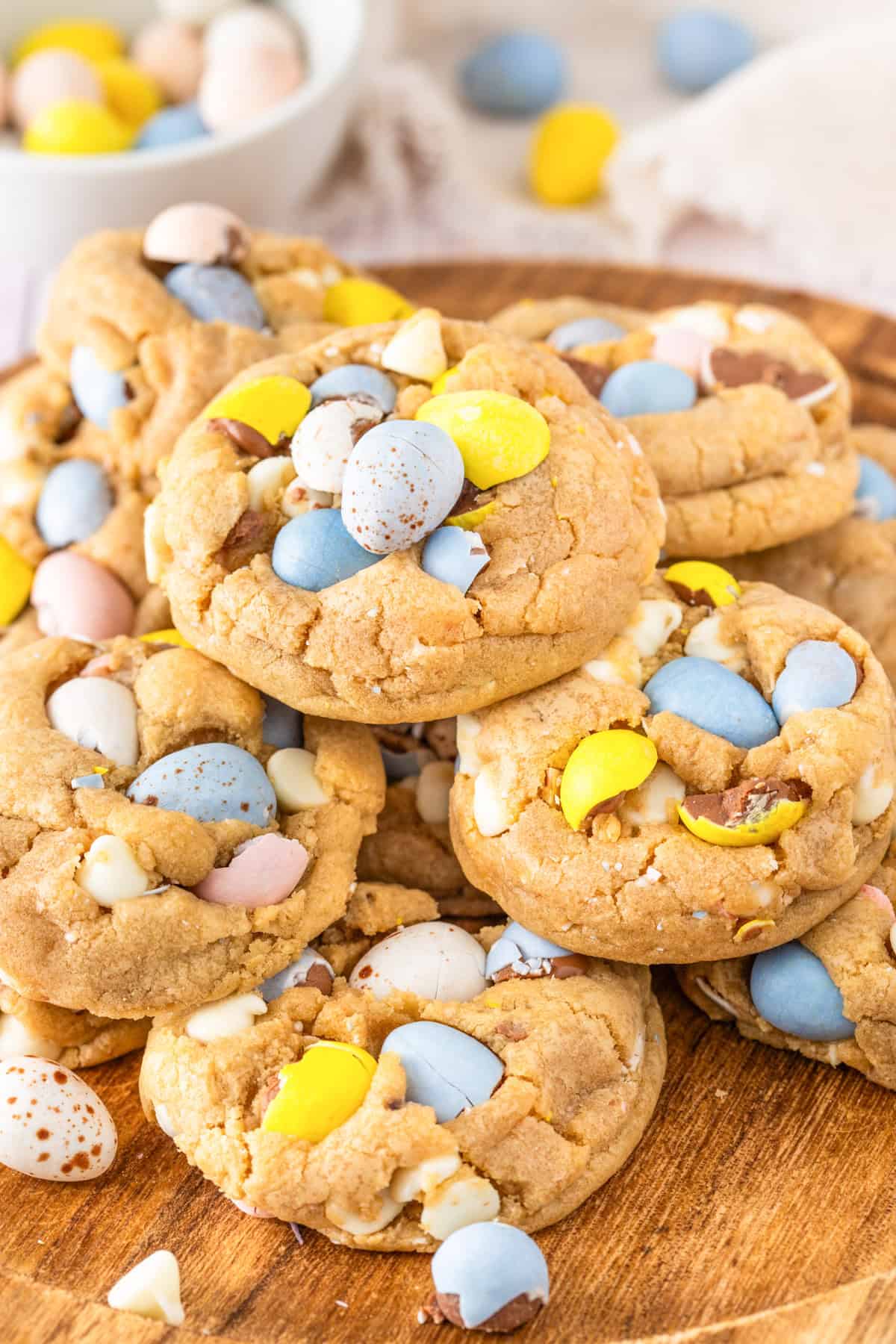 A stack of Mini Egg Chocolate Chip Cookies with pastel-colored candy-coated chocolate eggs and white chocolate chips on a wooden plate.