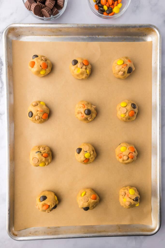A baking sheet with parchment paper holds twelve evenly spaced Reese’s Pieces peanut butter cookie dough balls.
