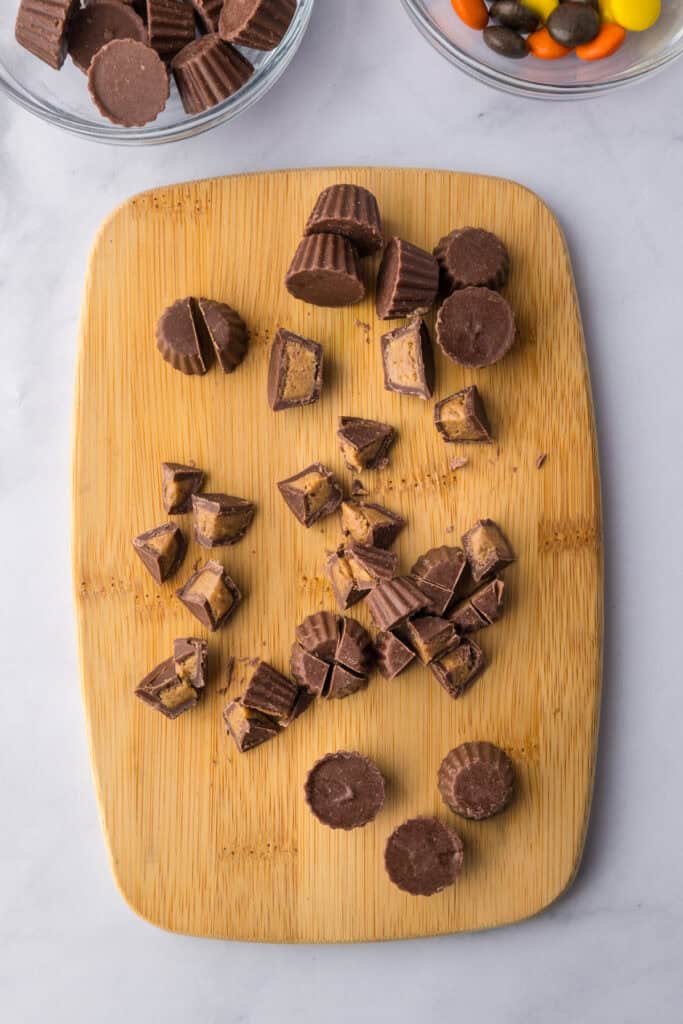A wooden cutting board with whole and chopped mini chocolate peanut butter cups for Reeses pieces peanut butter cookies.