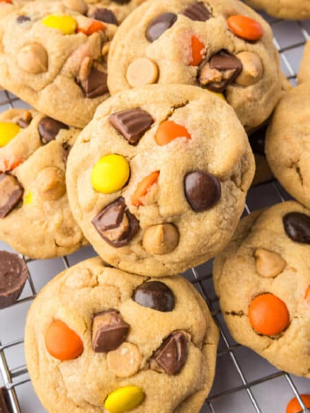 A close-up of Reeses pieces peanut butter cookies with chocolate chunks and colorful candy pieces, stacked on a wire cooling rack.