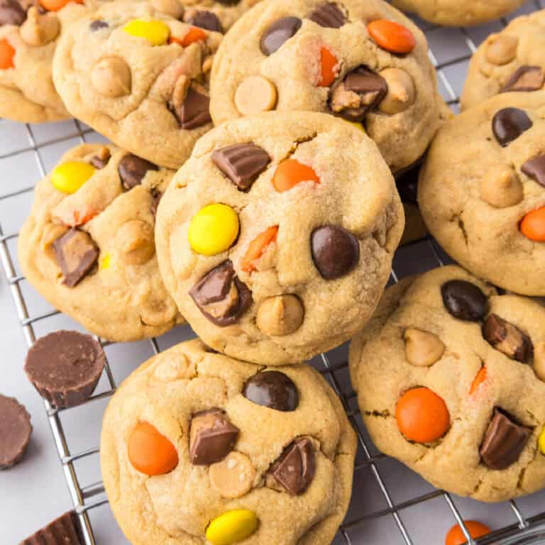 A close-up of Reeses pieces peanut butter cookies with chocolate chunks and colorful candy pieces, stacked on a wire cooling rack.