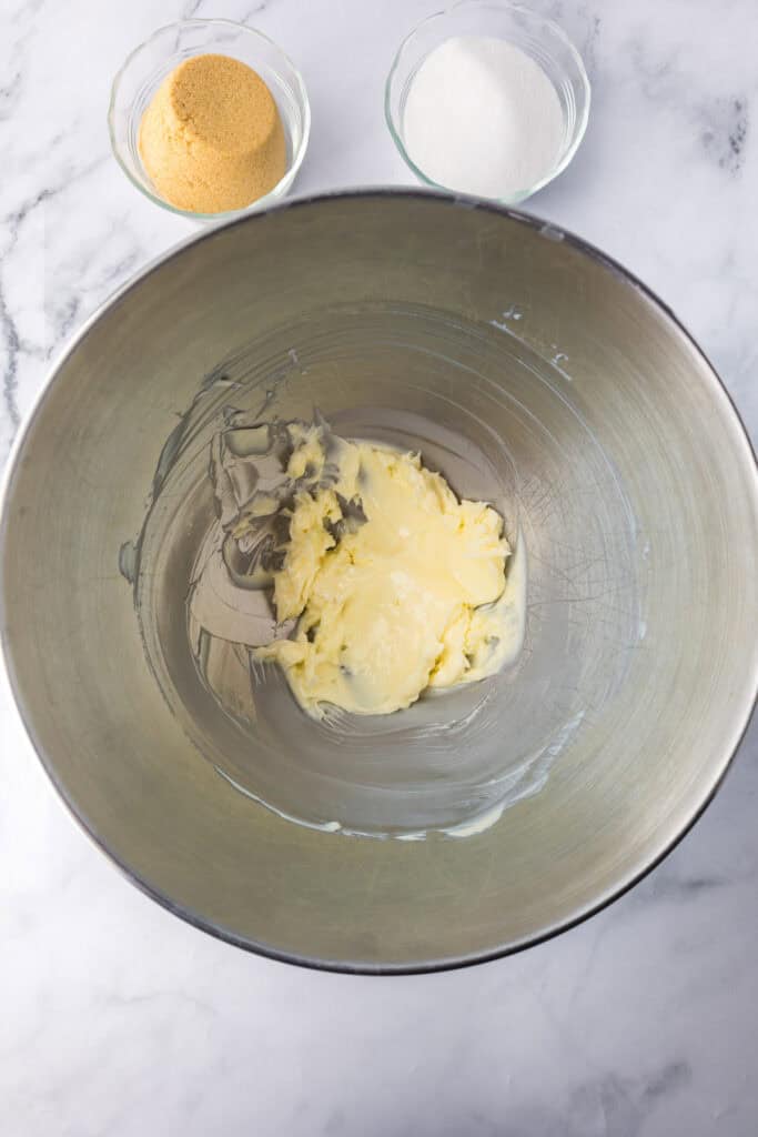 A mixing bowl with creamed butter next to small bowls of white sugar and brown sugar for Reeses pieces peanut butter cookies.