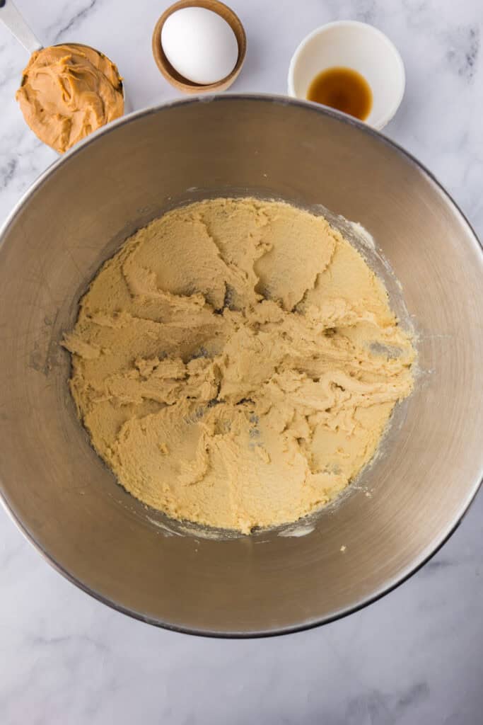 A metal mixing bowl with creamed butter and sugar for Reeses pieces peanut butter cookies with peanut butter, vanilla extract and an egg nearby.