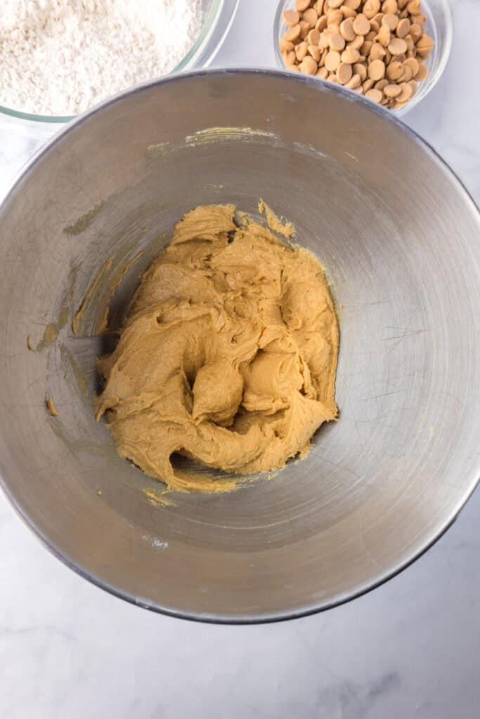 A mixing bowl with mixed wet ingredients for Reeses pieces peanut butter cookies with dry flour mixture and peanut butter chips nearby.