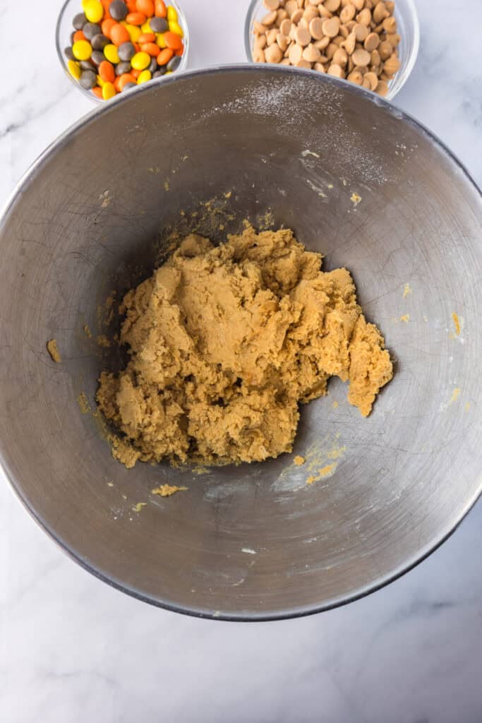 A metal mixing bowl contains cookie dough for Reeses Pieces peanut butter cookies next to a bowl of peanut butter chips and a bowl of Reeses pieces.