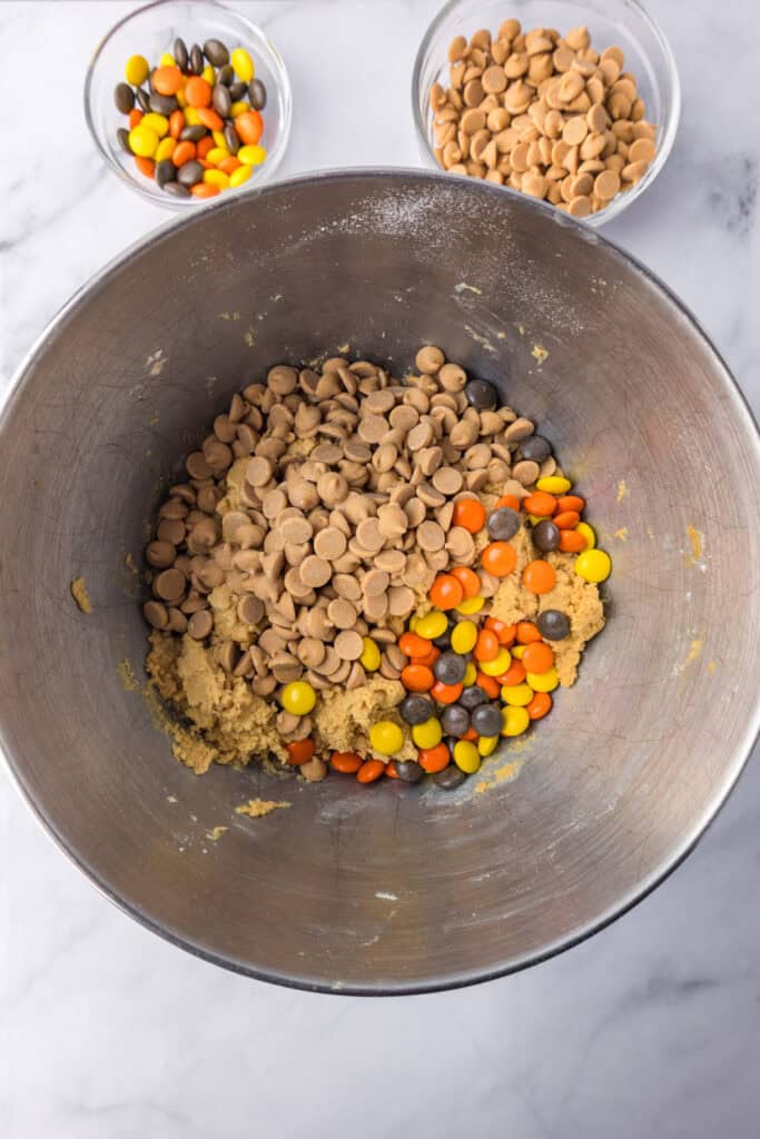 A metal mixing bowl holds cookie dough being mixed with peanut butter chips and Reese's pieces for Reeses pieces peanut butter cookies with bowls of more peanut butter candies nearby.