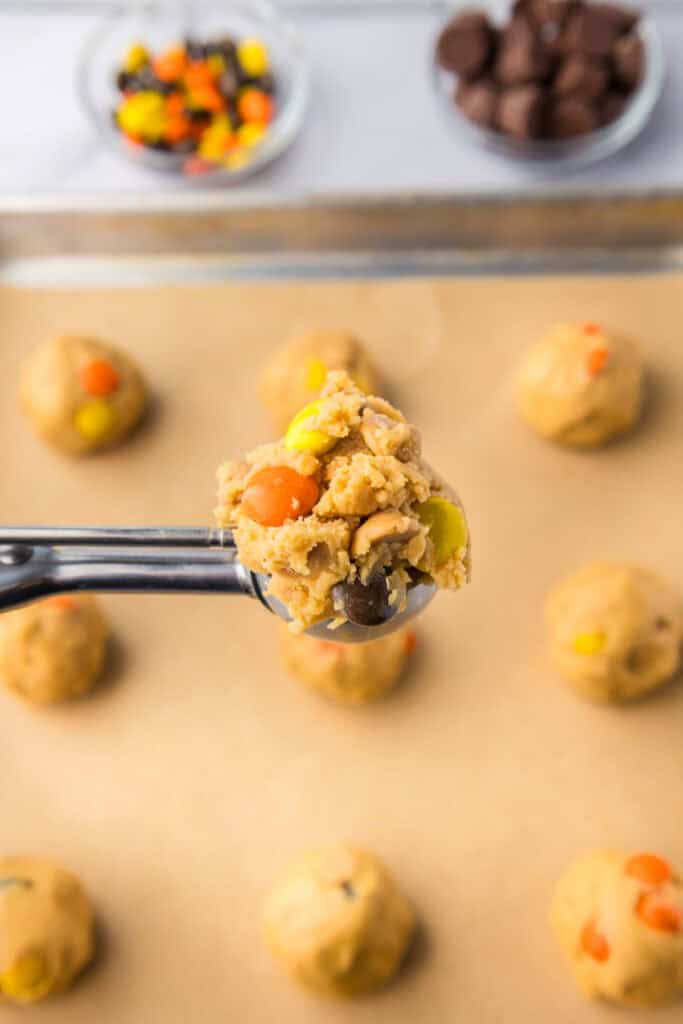 A cookie dough scoop holds a ball of Reeses pieces peanut butter cookies dough with more on a baking sheet in the background.