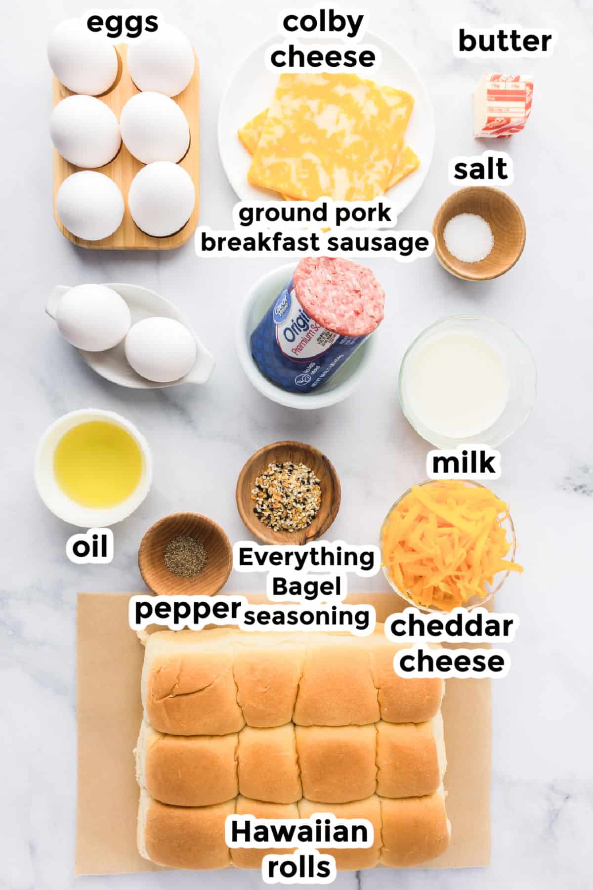 Ingredients for breakfast sliders are arranged on a counter, featuring eggs, cheeses, butter, sausage, milk, oil, spices, Hawaiian rolls, and seasoning.