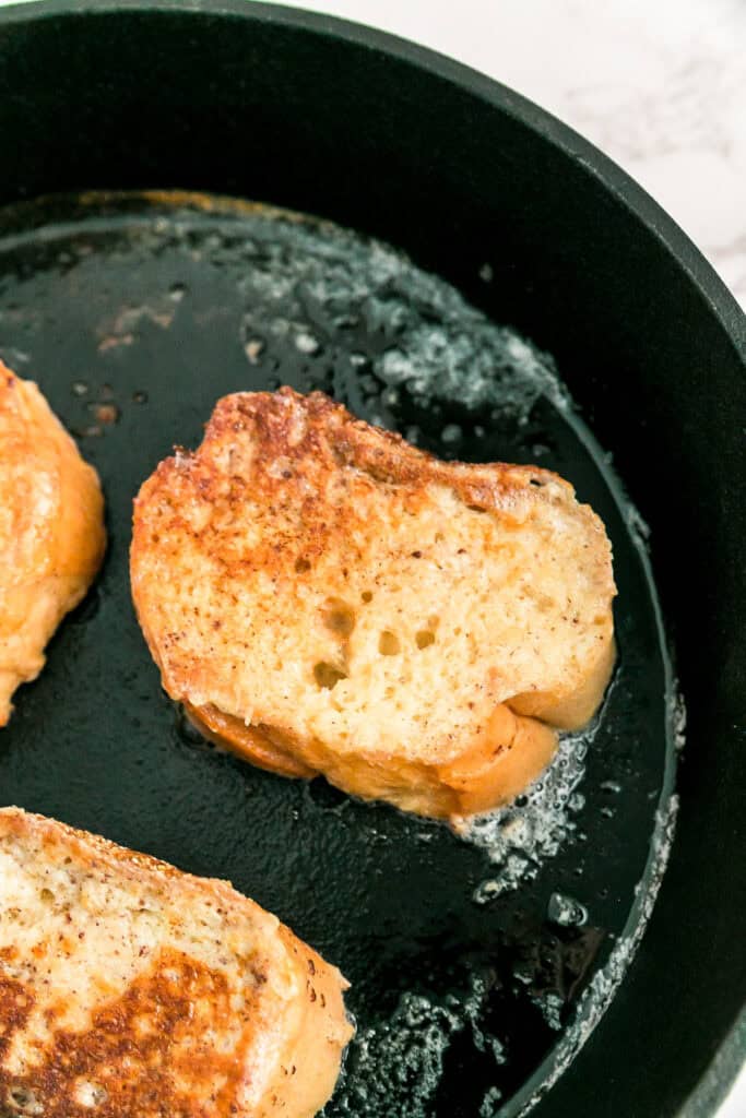 Slices of french bread french toast being cooked to a golden brown in a black skillet with melted butter.