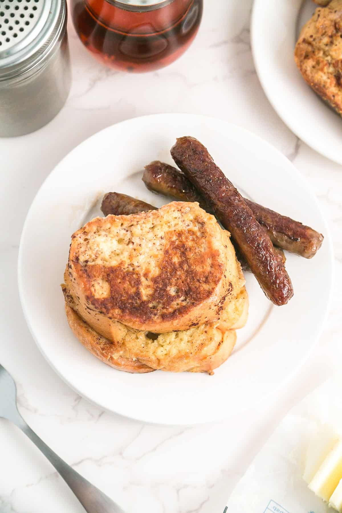 A white plate with two slices of French Bread French toast and three breakfast sausages, set on a marble surface with syrup, a shaker, and butter nearby.