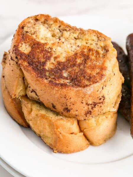 Two slices of golden brown French bread French toast stacked on a white plate next to two sausage links.