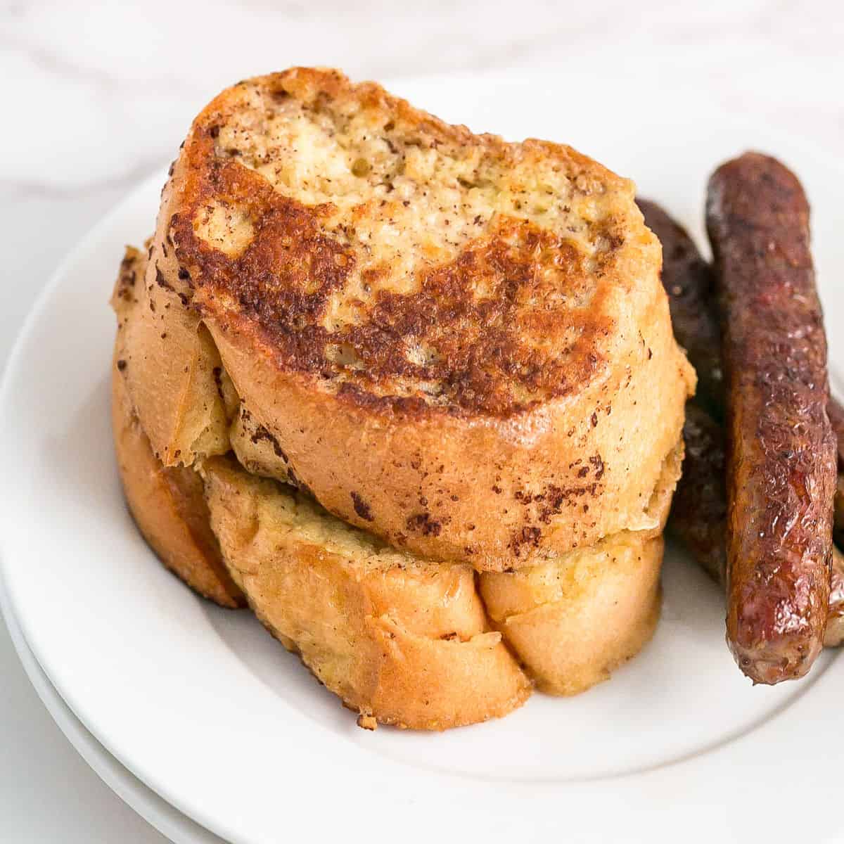 Two slices of golden brown French bread French toast stacked on a white plate next to two sausage links.