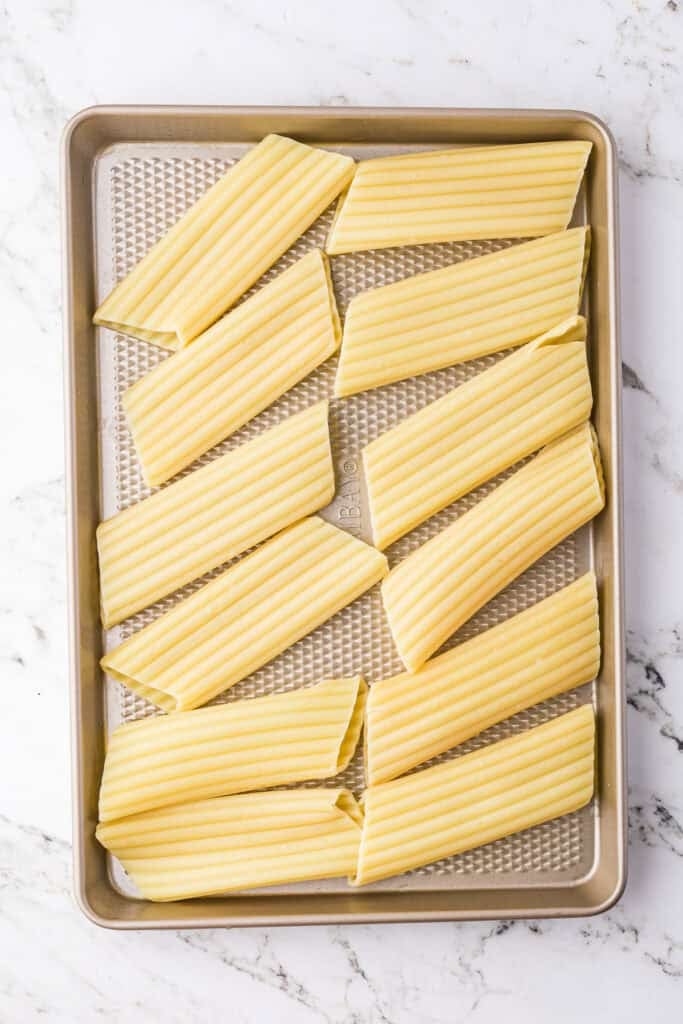 Cooked manicotti pasta tubes in rows on a metal baking sheet for making manicotti with sausage.
