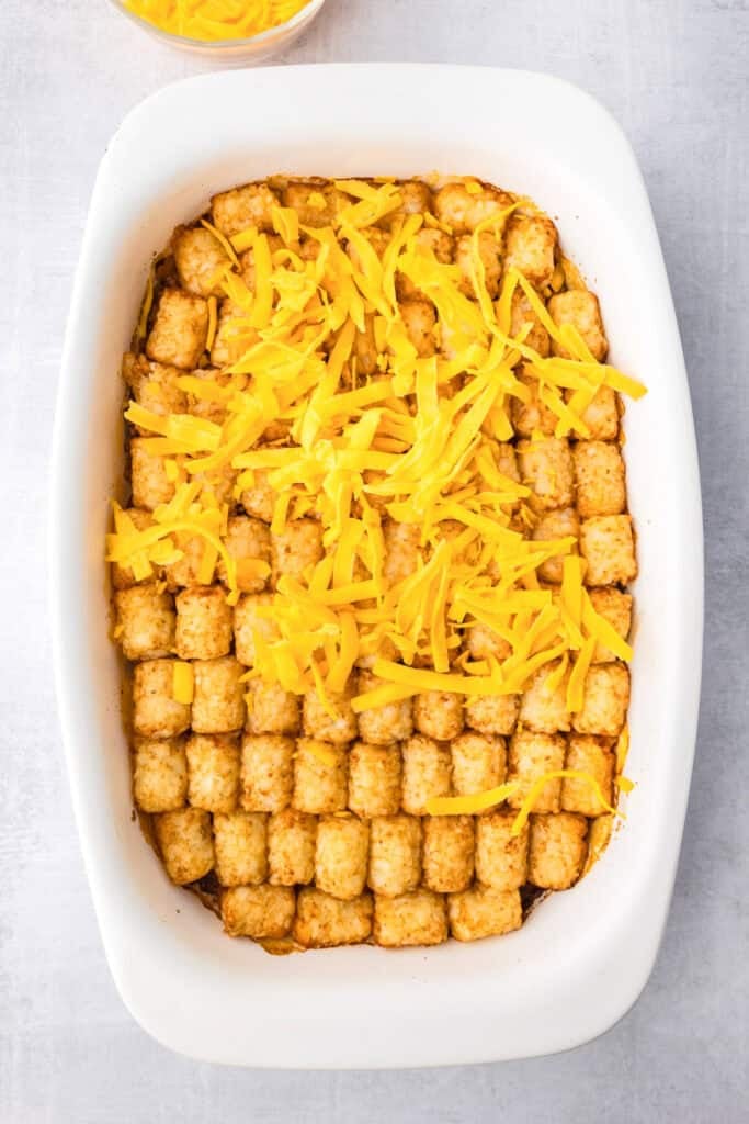 A white baking dish filled with sloppy joe tater tot casserole being topped with shredded cheddar cheese.