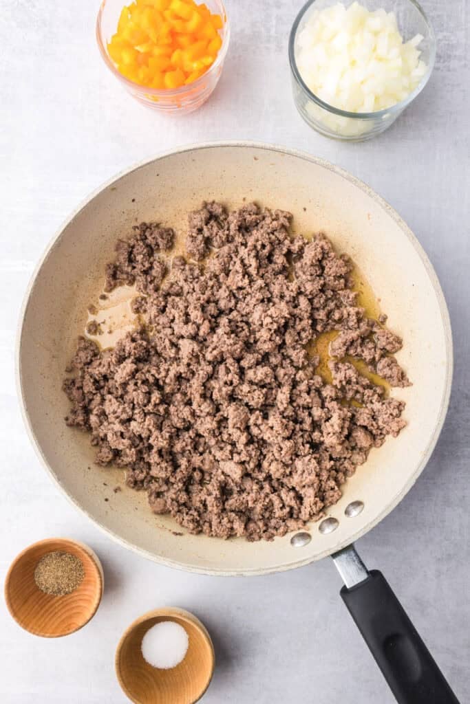 A skillet with cooked ground beef, surrounded by bowls of chopped onions, chopped bell peppers and spices for tater tot casserole.