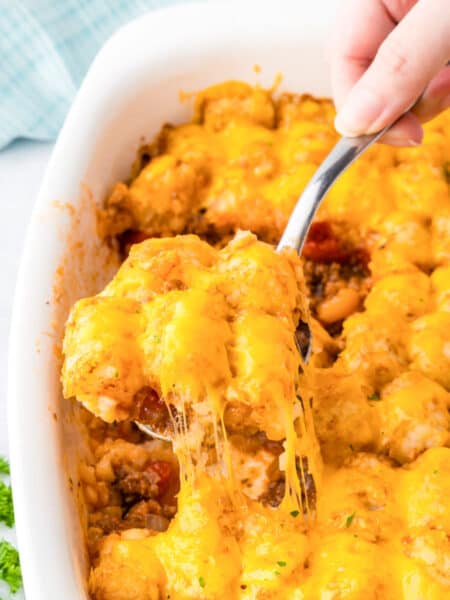 A hand scoops a serving of cheesy sloppy joe tater tot casserole from a white baking dish, with melted cheese stretching from the dish.
