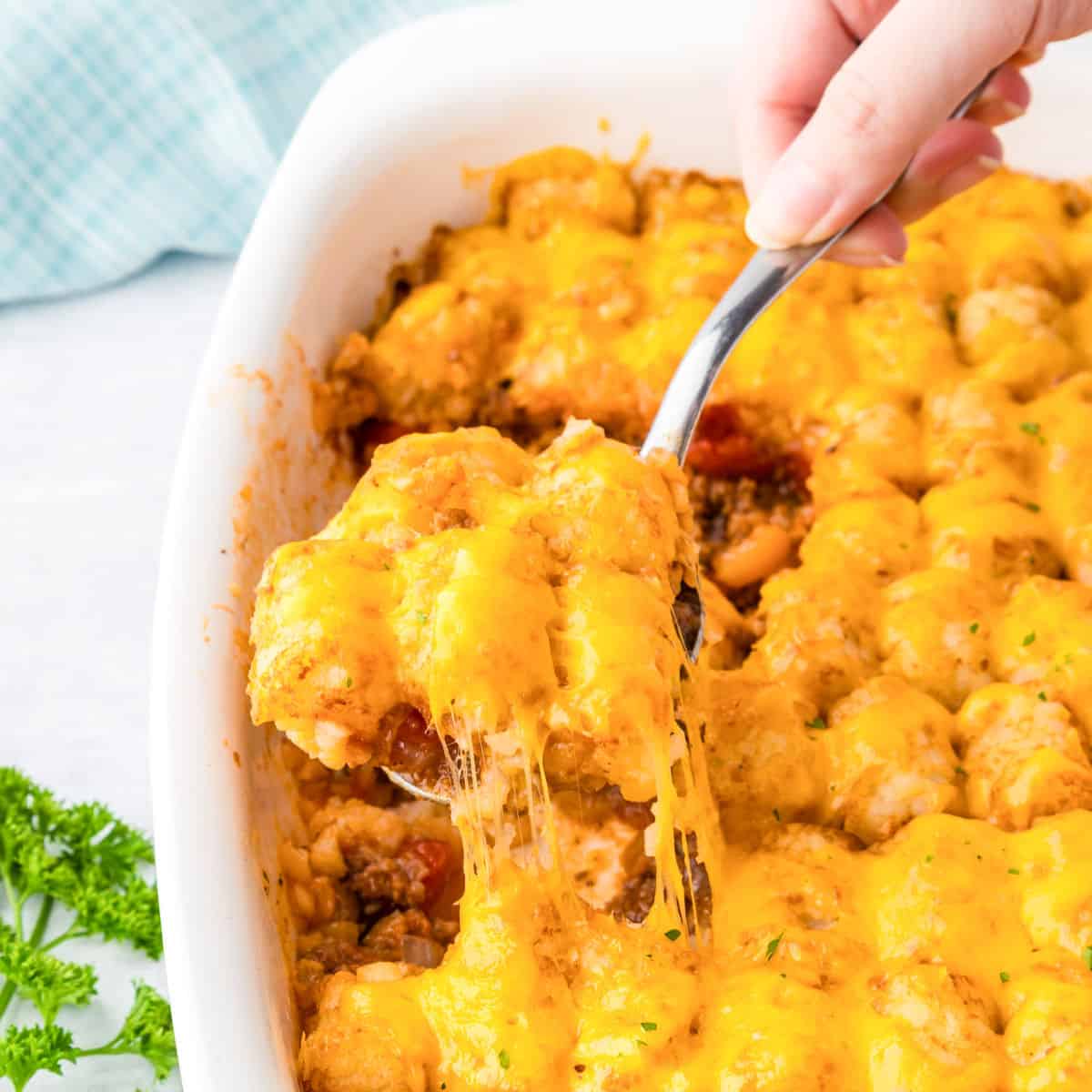 A hand scoops a serving of cheesy sloppy joe tater tot casserole from a white baking dish, with melted cheese stretching from the dish.
