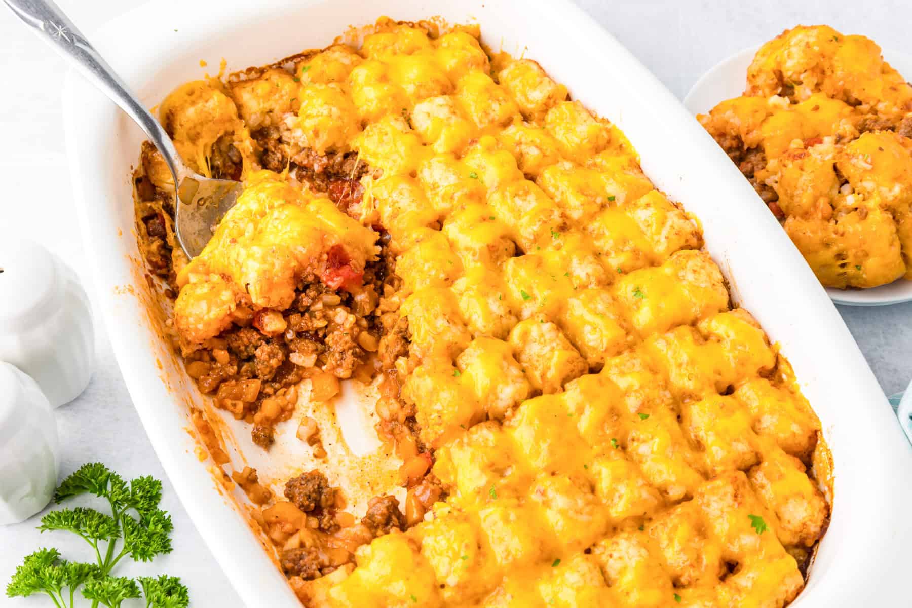 A casserole dish filled with sloppy joe tater tot casserole, topped with melted cheese, with a serving spoon scooping.