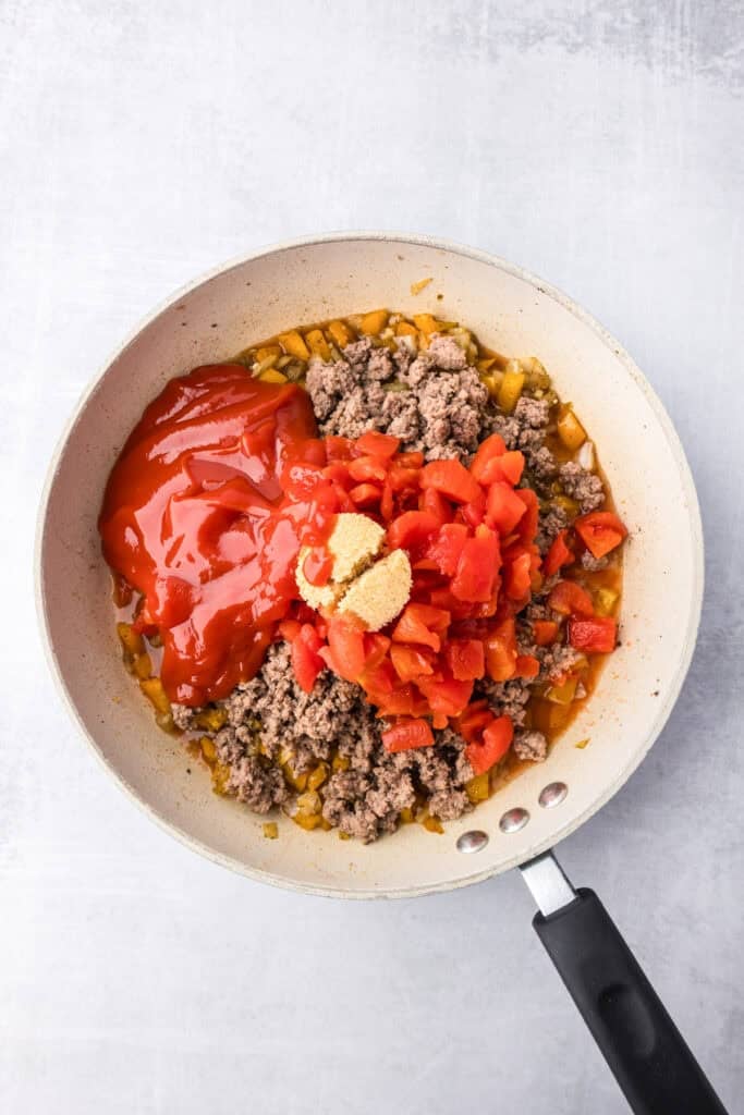 A frying pan containing cooked ground beef, chopped onions, and bell peppers being topped with tomatos and ingredients for the sauce of the sloppy joe tater tot casserole.