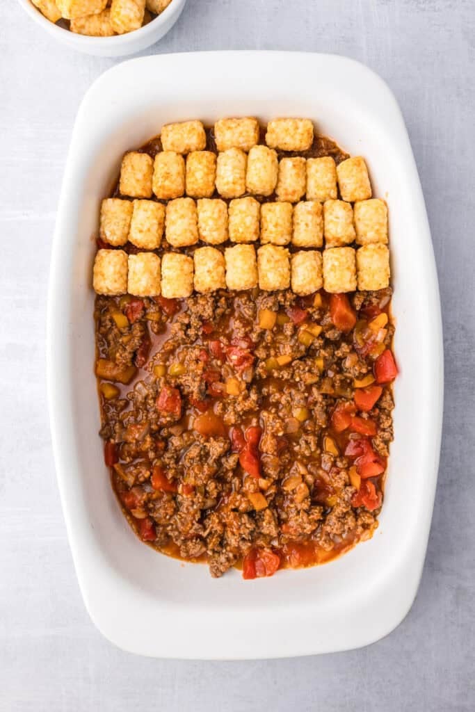 A white casserole dish filled with sloppy joe tater tot casserole with tater tots being lined up in rows.