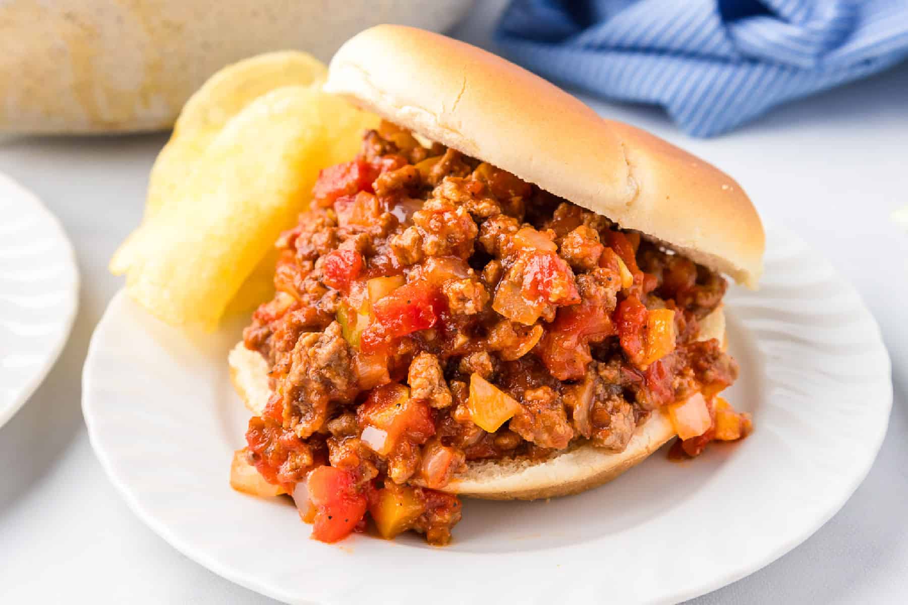 A sloppy joe sandwich filled with ground beef, tomatoes, and onions sits on a white plate next to a few potato chips.