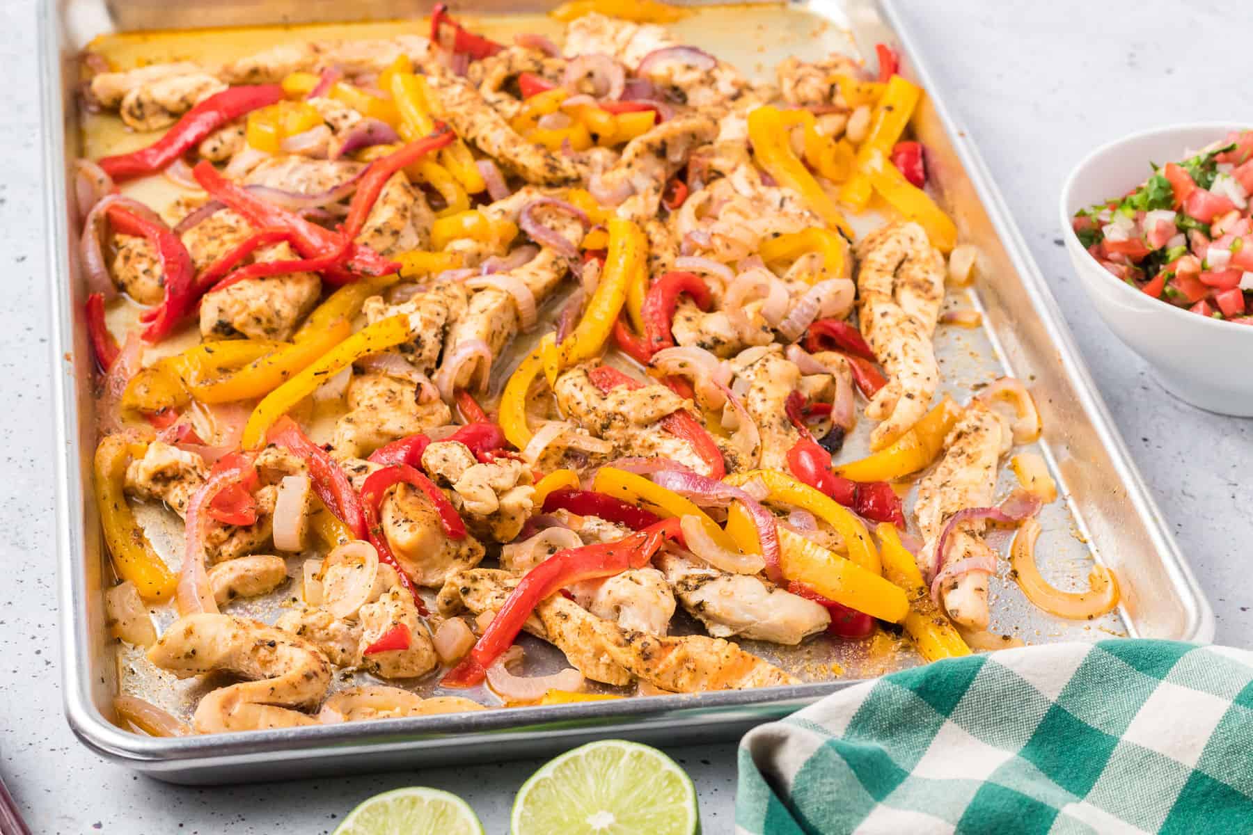 Sheet pan chicken fajitas with cooked chicken strips, red and yellow bell peppers, and onions; served with salsa, lime halves, and a green checkered cloth nearby.