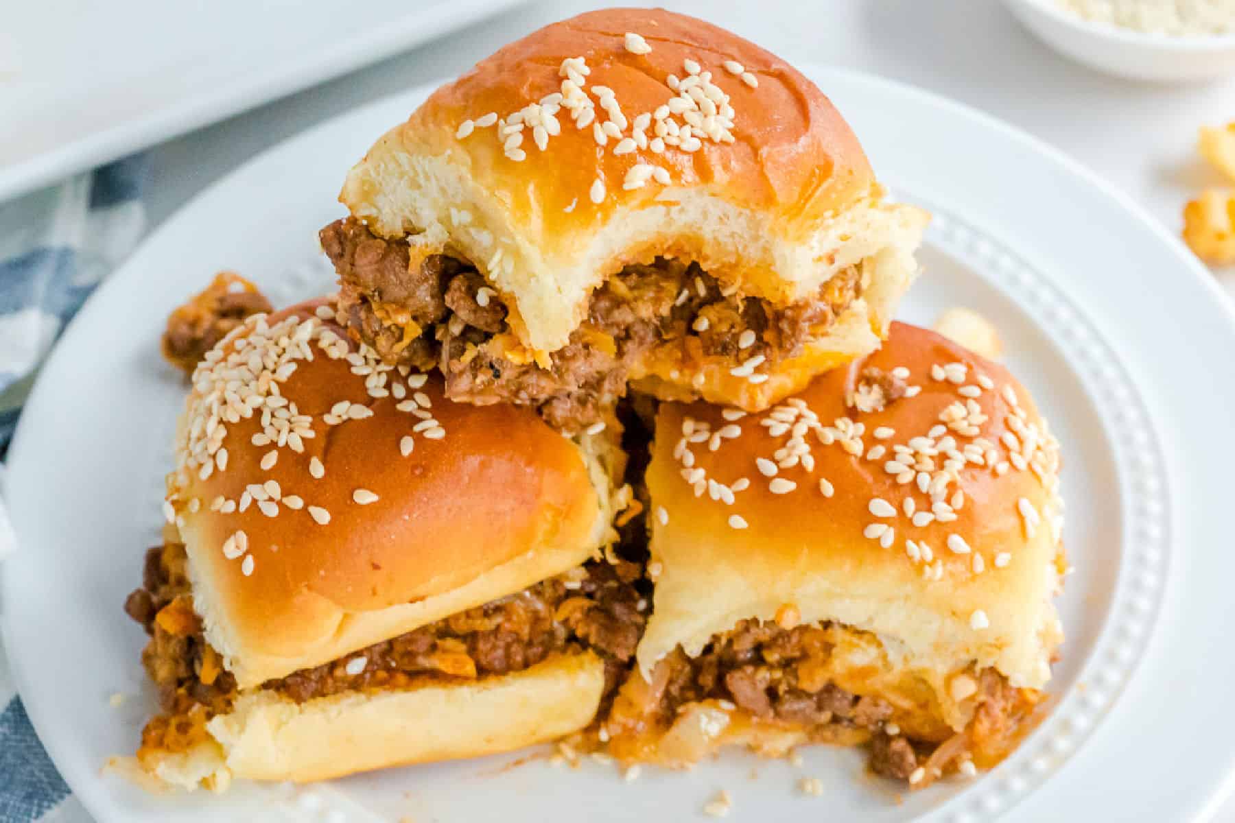Three sloppy joe sliders with sesame seed buns are stacked on a white plate, one slider missing a bite.