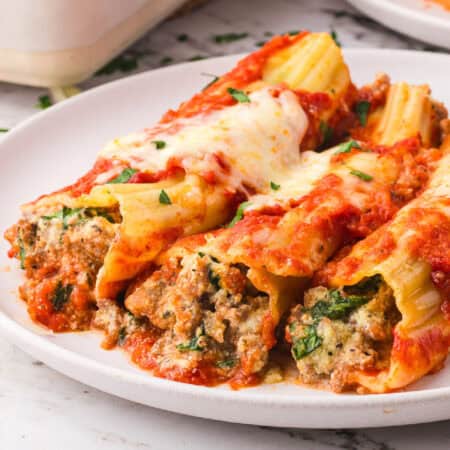 Three stuffed manicotti pasta shells stuffed with sausage filling and topped with melted cheese and tomato sauce are served on a white plate.