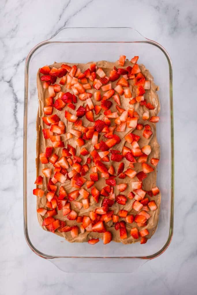 A glass baking dish filled with chocolate pudding mixture over graham crackers covered in a layer of diced strawberries for strawberry chocolate eclair cake.
