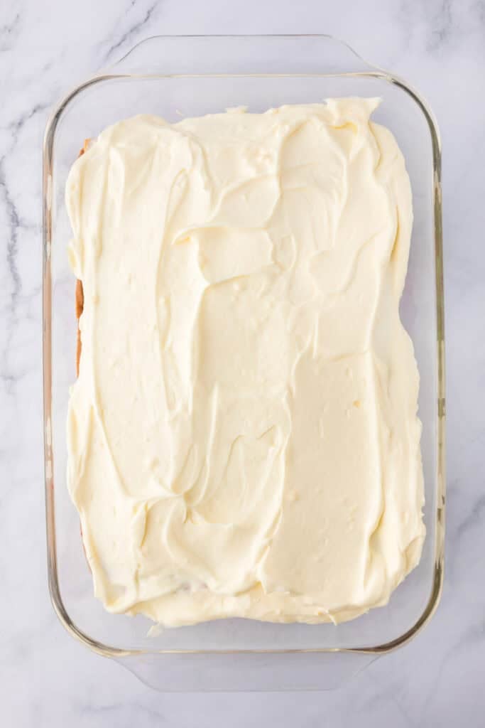 A rectangular glass dish filled with cheesecake pudding mixture layered layered completly over the other layers for strawberry chocolate eclair cake.
