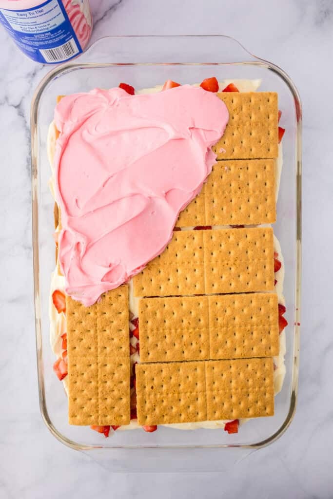 A glass dish with layers of graham crackers being topped with strawberry frosting over the otther layers for strawberry chocolate eclair cake.